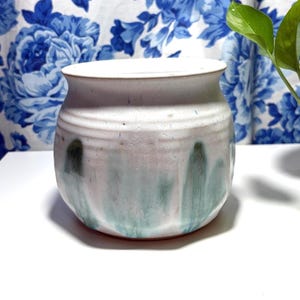May include: A handmade ceramic pot with a wide opening and a rounded body. The pot is primarily white with streaks of light blue and green. The pot is sitting on a white surface.