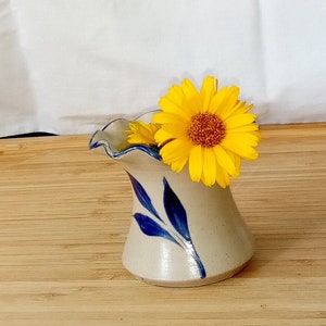 May include: A small, white ceramic vase with blue leaf designs, holding a single yellow flower with a brown center.