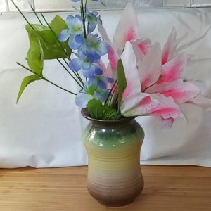 May include: A brown and green ceramic vase with a light yellow band around the middle. The vase is filled with pink and white lilies and blue delphiniums.