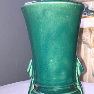 May include: A teal green ceramic vase with a flared opening and a rounded base. The vase has two sculpted handles on either side of the base.