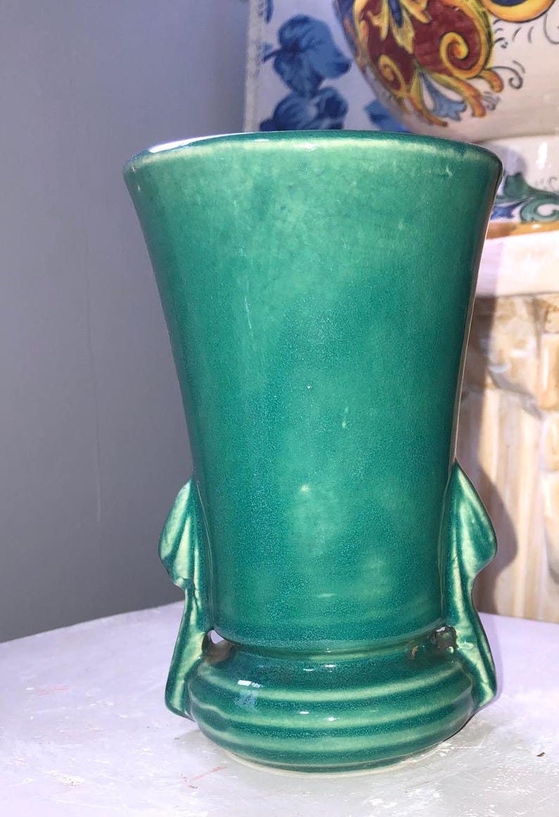 May include: A teal green ceramic vase with a flared opening and a rounded base. The vase has two sculpted handles on either side of the base.