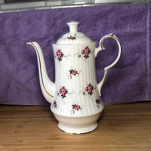 May include: A white ceramic teapot with a gold trim and a floral pattern of pink roses. The teapot has a lid and a handle.