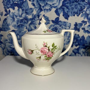 May include: Cream-colored porcelain teapot with a lid, handle, and spout. The teapot is decorated with pink roses and green leaves. Gold trim accents the rim and base. The teapot sits on a white surface against a blue floral background.