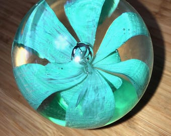 May include: A clear glass paperweight with a swirling, turquoise blue design inside. The paperweight is round and has a smooth, polished surface.