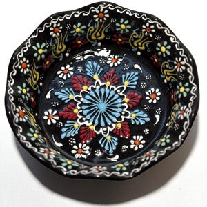 May include: A black ceramic bowl with a scalloped edge, adorned with a vibrant floral pattern. The design features a central blue flower surrounded by red, yellow, and white accents. The rim is decorated with a white, wavy border and small flowers.