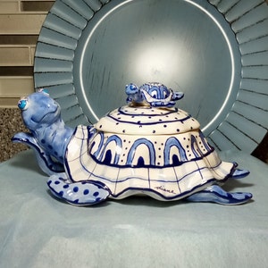 May include: A blue and white ceramic turtle with a smaller turtle on top. The larger turtle has a blue and white patterned shell and the smaller turtle has a blue shell with white dots. The larger turtle has the word "Diane" written on the bottom.