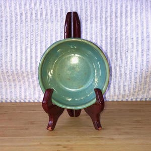 May include: A teal-colored ceramic bowl displayed on a dark wood stand. The bowl has a rounded shape with a slightly raised rim. The stand is a dark reddish-brown color and has a three-legged design. The background features a striped fabric.