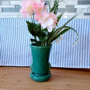 May include: A green ceramic vase with a unique design and three legs, holding a bouquet of pink and white flowers.