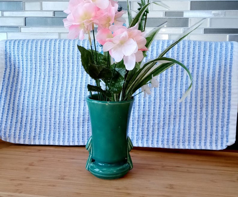 May include: A green ceramic vase with a unique design and three legs, holding a bouquet of pink and white flowers.