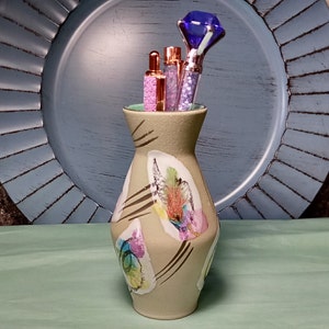 May include: A beige ceramic vase with a geometric design and colorful abstract patterns. The vase is filled with three pens and a blue jeweled pen topper.