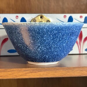 May include: A blue speckled ceramic bowl with a white base. The bowl has a rounded shape and a speckled pattern in shades of blue and white. The bowl is sitting on a wooden shelf.