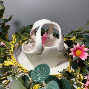 May include: A white ceramic basket with a handle, shaped like a basket with a pink interior. The basket is surrounded by green leaves and yellow flowers.