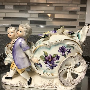 May include: A porcelain figurine of two children pushing a carriage decorated with purple and white flowers. The carriage has a single wheel and is detailed with gold accents.