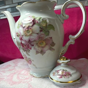 May include: White porcelain coffee pot with a floral design featuring pink and white roses and green leaves. The pot has a curved handle and a lid with a gold trim. The rim of the pot is also trimmed in gold.