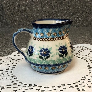 May include: A small blue and white ceramic creamer with a floral pattern. The creamer has a brown handle and a spout.