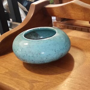 May include: A round, turquoise ceramic bowl with a speckled texture and a dark interior. The bowl is placed on a wooden surface, with wooden furniture visible in the background. The bowl has a glossy finish.