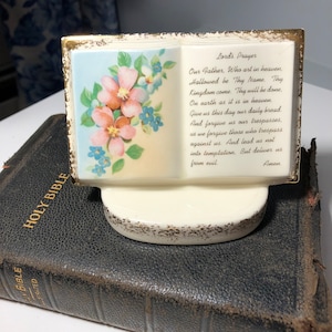 May include: A white ceramic bookend shaped like an open book with a gold trim. The bookend features a floral design on the front cover and the Lord's Prayer printed on the back cover.