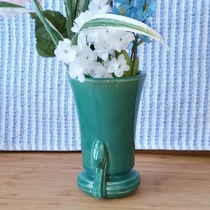 May include: A green ceramic vase with a unique design and a flared opening. The vase is filled with white and blue flowers.
