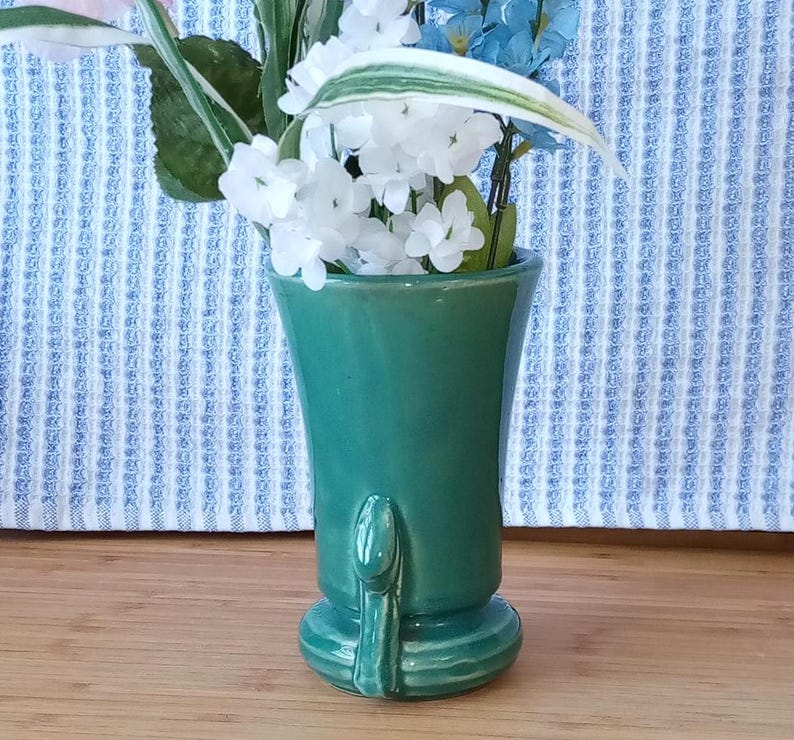 May include: A green ceramic vase with a unique design and a flared opening. The vase is filled with white and blue flowers.