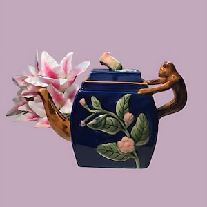 May include: A blue ceramic teapot with a monkey handle and floral design. The teapot has a lid with a flower on top. The teapot is decorated with pink flowers and green leaves.