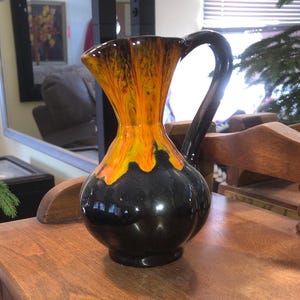 May include: A ceramic pitcher with a black base and handle, and a vibrant orange and yellow top. The pitcher has a glossy finish and a curved handle. It is sitting on a wooden surface.