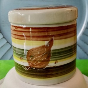 May include: A small, ceramic jar with a lid, featuring horizontal stripes in brown, yellow, and green. A brown leaf design is painted on the side. The jar is likely for kitchen or home decor.