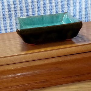 May include: A small, rectangular ceramic bonsai pot with a teal glaze and a dark brown rim. The pot has four small feet and is sitting on a wooden surface.