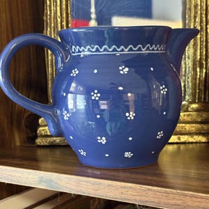 May include: A cobalt blue ceramic pitcher with a handle and spout. The pitcher is decorated with white floral and dot patterns, and a white wavy line near the rim. The pitcher is sitting on a wooden shelf.
