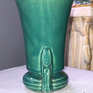 May include: A green ceramic vase with a flared top and a sculpted base. The vase has a smooth, glossy finish.