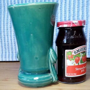 May include: A teal green ceramic vase with a rounded body and two handles. The vase is sitting on a wooden surface next to a glass jar of Smucker's strawberry jelly.
