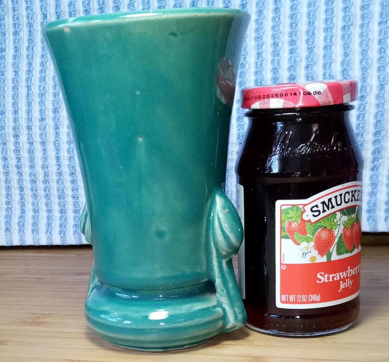 May include: A teal green ceramic vase with a rounded body and two handles. The vase is sitting on a wooden surface next to a glass jar of Smucker's strawberry jelly.