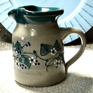 May include: A small, cream-colored ceramic creamer with a green rim and a leafy vine design in green, white, and blue.