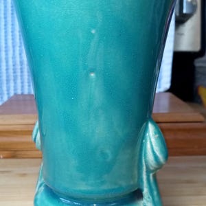 May include: A teal blue ceramic vase with a flared top and a rounded base. The vase has two sculpted handles on the sides.