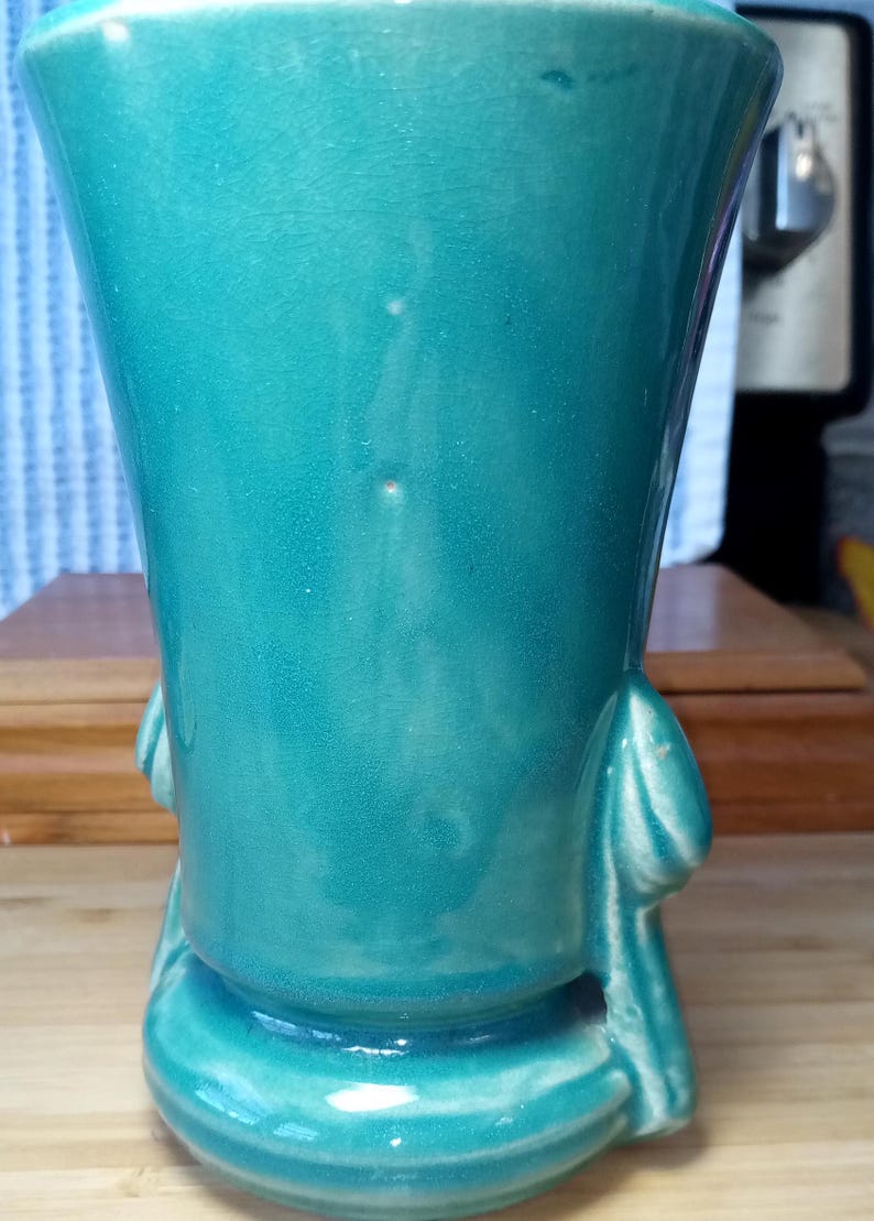 May include: A teal blue ceramic vase with a flared top and a rounded base. The vase has two sculpted handles on the sides.