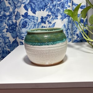 May include: A handcrafted ceramic pot with a white speckled base and a turquoise and green glazed top. The small, round pot is suitable for plants or decorative use. The pot is sitting on a white surface.