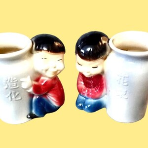 May include: Two white ceramic vases with a Chinese boy figurine on each side. The vases have Chinese characters on them. The boy on the left is holding a vase and the boy on the right is holding a vase.