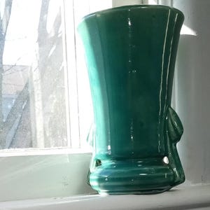 May include: A teal green ceramic vase with a wide mouth and a flared base. The vase has two handles that are shaped like leaves.