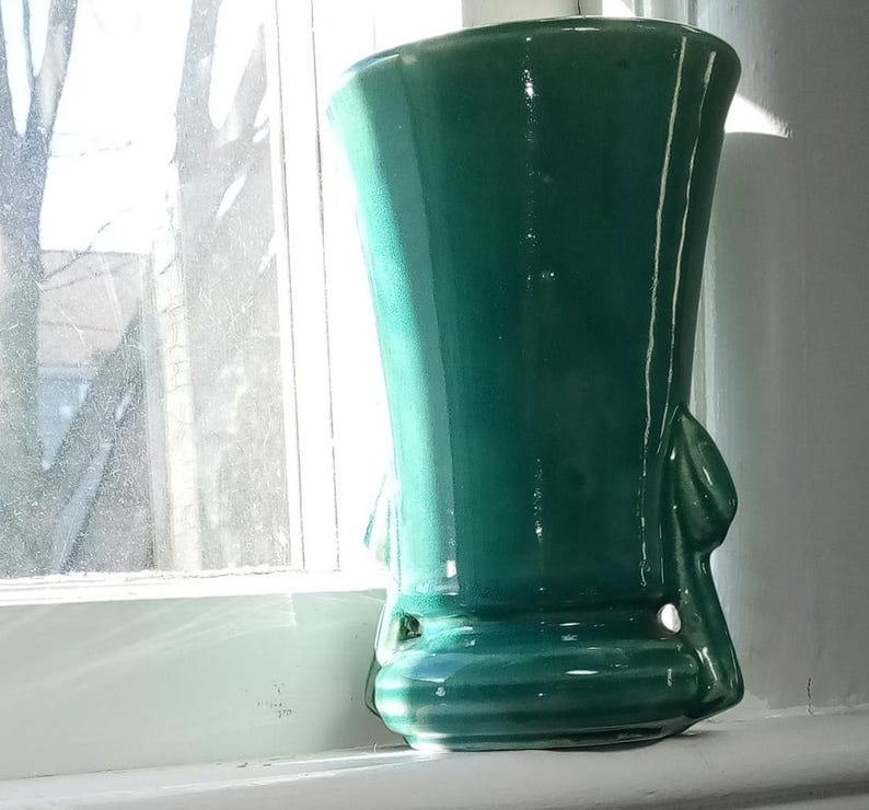 May include: A teal green ceramic vase with a wide mouth and a flared base. The vase has two handles that are shaped like leaves.