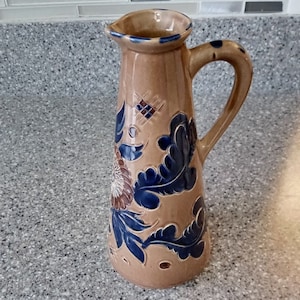 May include: Tan ceramic pitcher with a handle and spout. The pitcher is decorated with blue and brown floral designs. The handle has blue dots. The pitcher is approximately 10 inches tall.