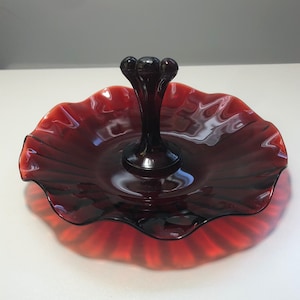 May include: A red glass candy dish with a fluted edge and a central pedestal. The dish has a scalloped rim and a raised center.