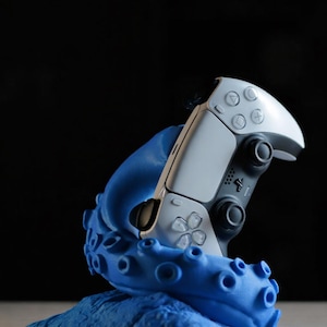 Könnte beinhalten: Ein weiß-schwarzer kabelloser Videospiel-Controller, der von einem leuchtend blauen, texturierten Krakenarm-Ständer gehalten wird. Der Controller verfügt über Standard-Richtungstasten, zwei Analogsticks und ein zentrales Touchpad auf der Basis.