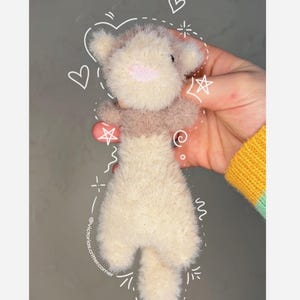 May include: A small, white and brown plush toy shaped like a dog. The toy has a pink nose and is being held in a hand. The background is a light gray wall with white doodles.