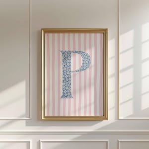 May include: A framed print with the letter 'P' in a blue floral design, set against a backdrop of pink and white vertical stripes. The artwork is encased in a gold-coloured frame, suitable for home decor.