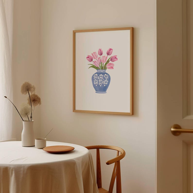 May include: A framed art print featuring a blue vase with white floral designs, holding pink tulips. The artwork is displayed on a cream-colored wall. The frame is made of light brown wood.