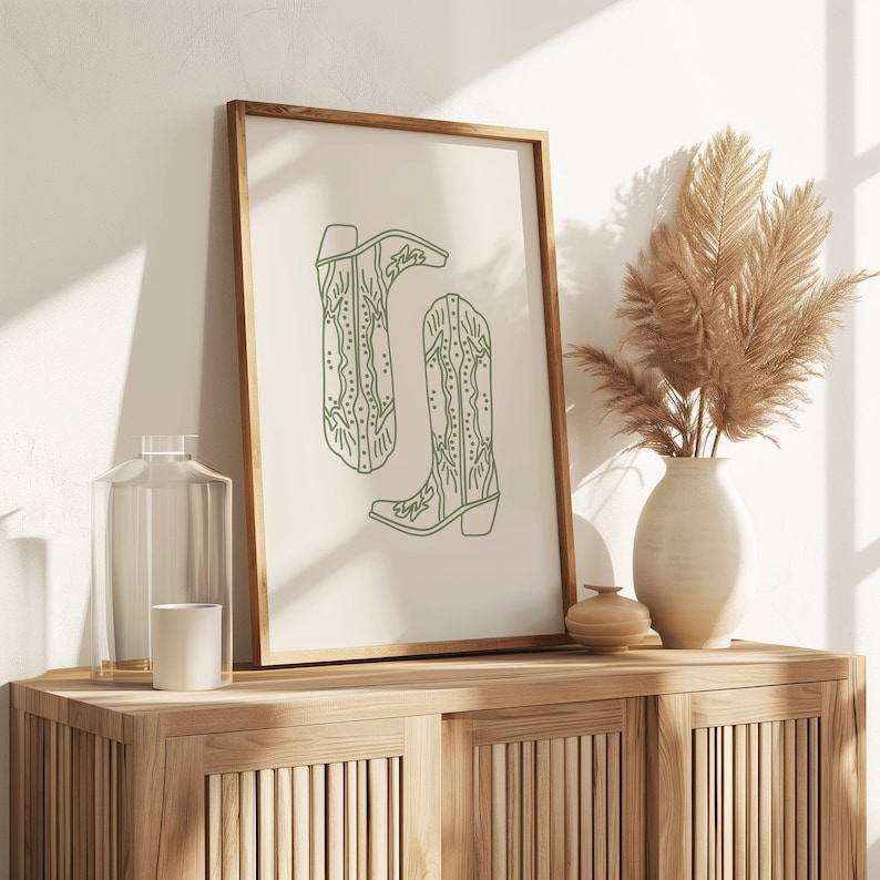 May include: A framed print with a minimalist line drawing of two cowboy boots in a soft green hue. The artwork is displayed on a wooden cabinet, alongside a vase with dried pampas grass, a glass bottle, and a candle.