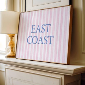 May include: A framed artwork with a pink and white striped pattern and the words "EAST COAST" in blue. The artwork has a light wooden frame and is displayed on a white mantelpiece. A gold lamp is to the left.
