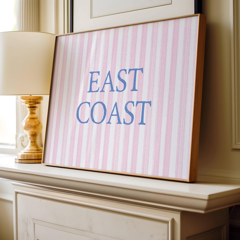 May include: A framed artwork with a pink and white striped pattern and the words "EAST COAST" in blue. The artwork has a light wooden frame and is displayed on a white mantelpiece. A gold lamp is to the left.