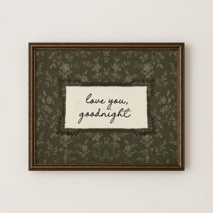 May include: A framed wall art piece featuring a dark olive green background with a subtle floral pattern. In the center, a cream-colored rectangular fabric patch displays the black cursive text "love you, goodnight" surrounded by a dark lace border.
