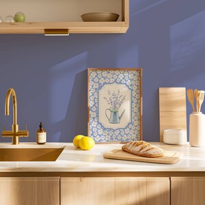 May include: A kitchen scene featuring a framed floral print with a watering can, a loaf of bread on a cutting board, and a gold faucet. The countertop has lemons and a bottle of soap. The background is a solid blue wall.