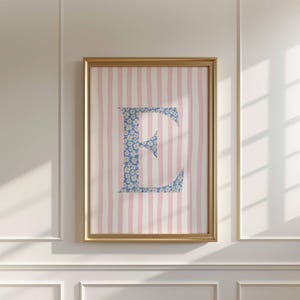Puede incluir: Obra de arte enmarcada con la letra "E" en un patrón floral azul. El fondo presenta rayas verticales rosas y blancas. El marco dorado añade un toque de elegancia a esta pieza decorativa, ideal para un dormitorio infantil.